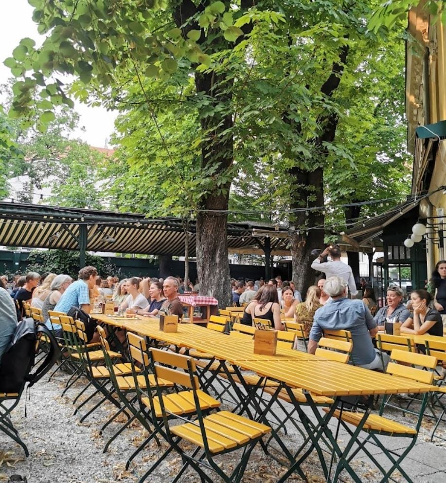 Fischerbräu beer garden in Vienna with yellow tables under trees, guests enjoying drinks and food
